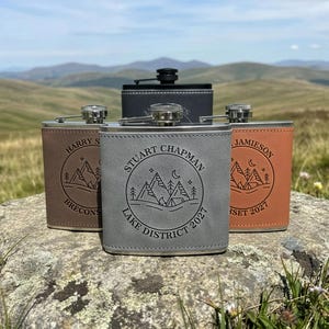 May include: Four rectangular metal flasks wrapped in leather-like material in grey, brown, tan, and black. Each flask features a circular engraving of a mountain landscape with trees, a crescent moon, and personalised text including names and dates.