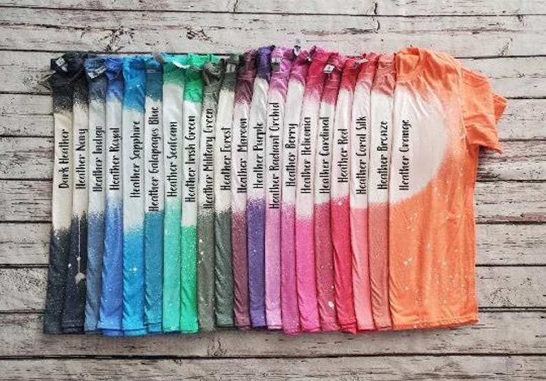 May include: A collection of colorful t-shirts with a bleached design. Each shirt is a different color, including dark heather, navy, indigo, royal, sapphire, and more. The shirts are arranged on a wooden surface.