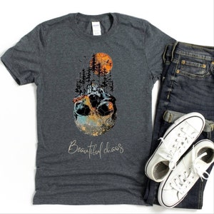 May include: A dark heather grey t-shirt with a graphic of a skull with a forest growing out of it. The graphic is surrounded by a colorful, abstract design. The text "Beautiful chaos" is printed below the graphic.