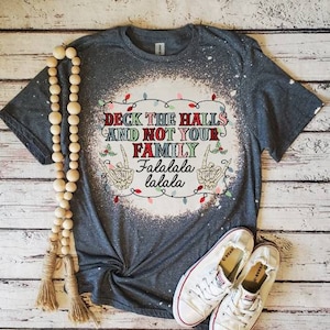 May include: A gray t-shirt with a bleach-splattered design featuring the text "Deck the Halls and Not Your Family Falalalala lalala" with a string of Christmas lights and two skeleton hands making the peace sign.