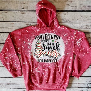 May include: Red hooded sweatshirt with a white bleach splattered design. The design features a Christmas tree and the text "Torn between lookin' like a snack and eatin' one".