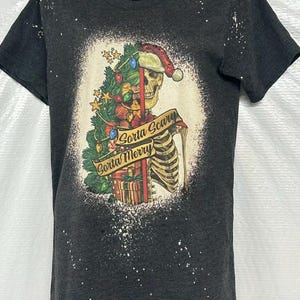 May include: Dark gray t-shirt featuring a skeleton wearing a Santa hat and holding a Christmas tree. The graphic includes the text "Gorta Scary Gorta Merry" on a banner. The shirt has a bleached, distressed look.