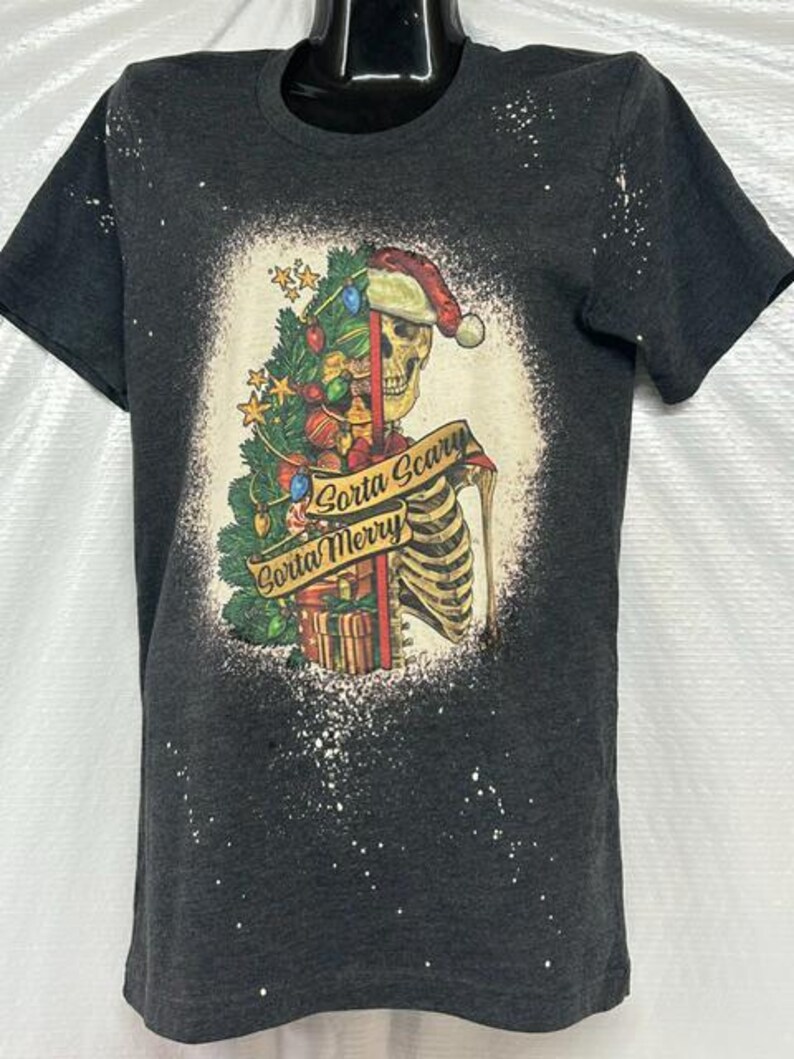 May include: Dark gray t-shirt featuring a skeleton wearing a Santa hat and holding a Christmas tree. The graphic includes the text "Gorta Scary Gorta Merry" on a banner. The shirt has a bleached, distressed look.