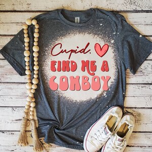 May include: A gray t-shirt with the words "Cupid. Find Me A Cowboy" in red and pink lettering. The shirt has a bleached design around the text and is paired with white sneakers and a wooden bead garland.
