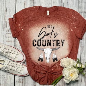 May include: A rust-colored t-shirt with a bleached design. The design features the text "This is God's Country" with a skull and crossbones and feathers.