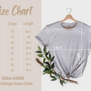 May include: A light gray Gildan 64000 Adult Softstyle Unisex T-Shirt hanging on a wooden hanger. The image includes a size chart with chest and length measurements in inches, and a sprig of greenery.