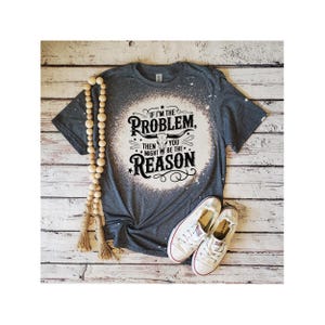 May include: A dark gray short-sleeve t-shirt with a bleached circular center featuring a black graphic of a longhorn skull and the text IF I'M THE PROBLEM, THEN YOU MIGHT BE THE REASON. A strand of wooden beads and white canvas sneakers are included.