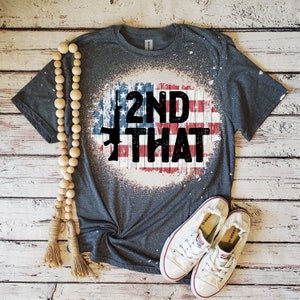 May include: A gray t-shirt with a distressed American flag design and the text "2ND THAT" printed in black. The flag design includes a stylized rifle.