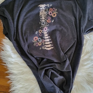 Pu&ograve; includere: T-shirt grigio scuro con un design della colonna vertebrale adornato da fiori colorati. La grafica include un'illustrazione dettagliata della colonna vertebrale con vari accenti floreali dai colori pastello. La maglietta &egrave; in materiale morbido.