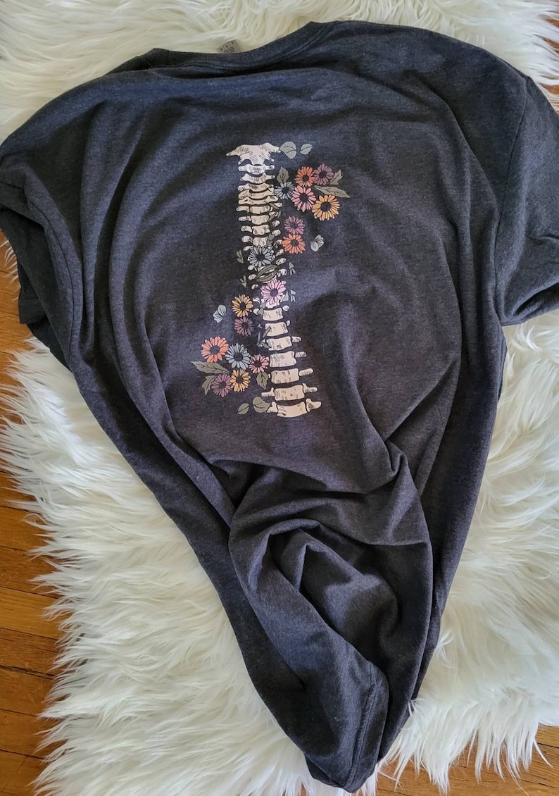 Pu&ograve; includere: T-shirt grigio scuro con un design della colonna vertebrale adornato da fiori colorati. La grafica include un'illustrazione dettagliata della colonna vertebrale con vari accenti floreali dai colori pastello. La maglietta &egrave; in materiale morbido.