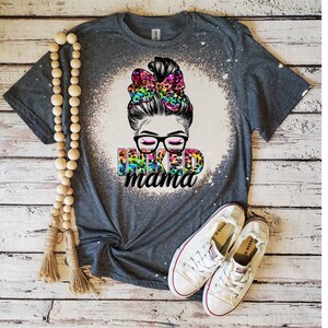 May include: A gray t-shirt with a bleach-splattered design featuring a woman with a messy bun, glasses, and a leopard print bow. The text "INKED mama" is printed in rainbow colors.