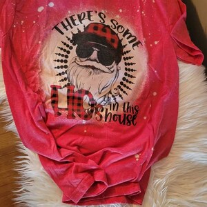 May include: Red t-shirt with a bleach-splattered design featuring a cartoon Santa Claus wearing sunglasses and a plaid hat. The text "There's some Ho in this house" is printed on the shirt.