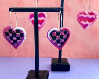 Op de afbeelding: Twee paar hartvormige oorbellen op een zwarte standaard. Het ene paar is roze en wit met een schaakbordpatroon, het andere is roze en wit met golvende lijnen.