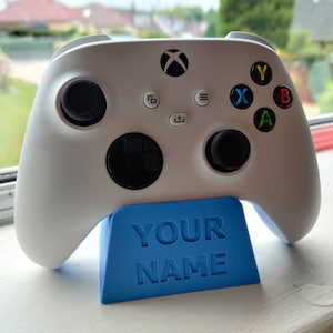 May include: A white wireless video game controller with black joysticks and colored action buttons, resting on a blue plastic stand featuring the embossed text YOUR NAME on the front face, designed for holding the controller upright.