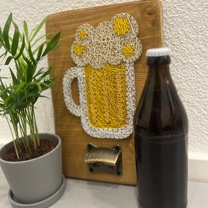 Könnte beinhalten: Ein Holzschild mit einem String-Art-Design eines Bierkruges mit Schaum oben drauf. Das Schild hat einen Flaschenöffner mit der Aufschrift "OPEN HERE" angebracht. Eine braune Glasflasche Bier steht vor dem Schild.
