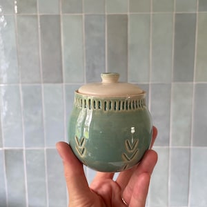May include: A small, handcrafted ceramic jar with a lid. The jar is a muted teal colour with a cream-coloured lid and a series of vertical cutouts near the top. Decorative arrow-like patterns are carved into the jar's surface.