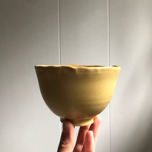 Puede incluir: Un cuenco de cerámica amarillo con un borde ligeramente irregular. El cuenco está hecho a mano y tiene un aspecto rústico y orgánico.