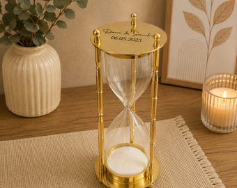 May include: A gold-toned metal hourglass with a clear glass bulb containing white sand. The top circular plate features the engraved text "Dani & Sandra" and the date "06.05.2023" in a black script font. The frame consists of four vertical pillars.