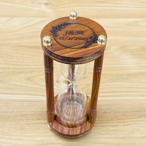 May include: A wooden hourglass with a clear glass center. The top and bottom are round, with gold-colored accents. The top has the initials "J&M" and the date "11/2/2023" engraved within a circular design. The hourglass is set on a wooden surface.