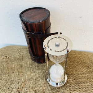May include: A silver hourglass with a bamboo-style stand and a round top inscribed with "GREATEST STAR CATCHER WORLD TOUR TOYOTA CENTER JANUARY 13, 2023". The hourglass is in a brown leather carrying case.