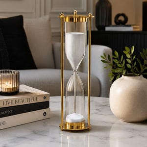 May include: A tall hourglass featuring a polished gold metal frame with vertical supports and circular end caps. The clear glass vessel contains fine white sand, with a portion currently flowing from the top bulb into the bottom bulb.