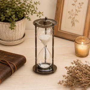 May include: A vintage-style hourglass with a bronze metal frame and clear glass bulbs. The hourglass contains white sand and is placed on a wooden surface. The top of the hourglass has the text "MEMORY OF TIME".