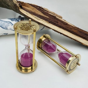 May include: Two brass hourglass sand timers with pink sand. The top of one hourglass has an engraved message: "Only  measure yourself by what is  at the end of every  affliction with love and  patience.  Best wishes,  Charlie."