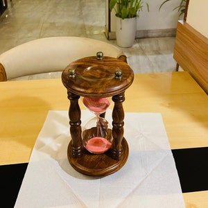 May include: A wooden hourglass with a dark brown finish and brass accents. The hourglass contains pink sand and has a classic, decorative design. The hourglass is a decorative item.