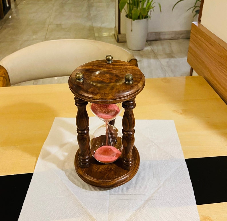 May include: A wooden hourglass with a dark brown finish and brass accents. The hourglass contains pink sand and has a classic, decorative design. The hourglass is a decorative item.