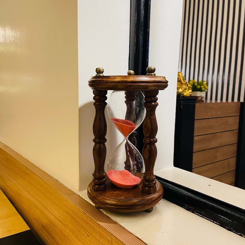 May include: A wooden hourglass with a dark brown finish. The hourglass has a clear glass chamber filled with red sand. The hourglass is approximately 6 inches tall and sits on a wooden base.