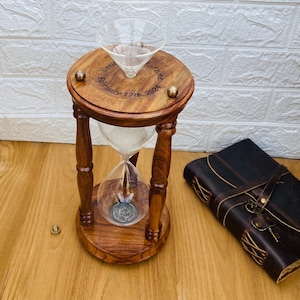 May include: A wooden hourglass with a clear glass center, filled with sand. The top of the hourglass has the inscription "Until February 17th, 2024." Brass accents adorn the wooden frame. A leather-bound journal with a key is next to the hourglass.