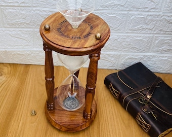 May include: A wooden hourglass with a clear glass center, filled with sand. The top of the hourglass has the inscription "Until February 17th, 2024." Brass accents adorn the wooden frame. A leather-bound journal with a key is next to the hourglass.