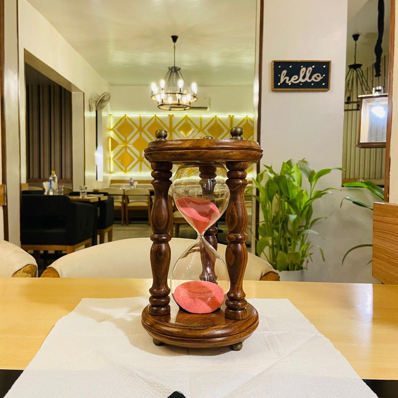 May include: A wooden hourglass with a dark brown finish, featuring a clear glass bulb filled with pink sand. The hourglass is set on a light-colored surface, with a white napkin underneath. The word "hello" is written on a small sign in the background.