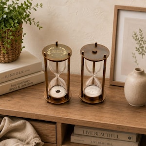 May include: Two hourglasses with antique brass-toned metal frames and clear glass bulbs containing white sand. The left hourglass features a circular top plate engraved with a gold-colored laurel wreath design and text. The frames have vertical supports.