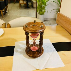 May include: A wooden hourglass with a dark brown finish and brass accents. The hourglass contains pink sand and is set on a wooden base. The hourglass is approximately 6 inches tall.