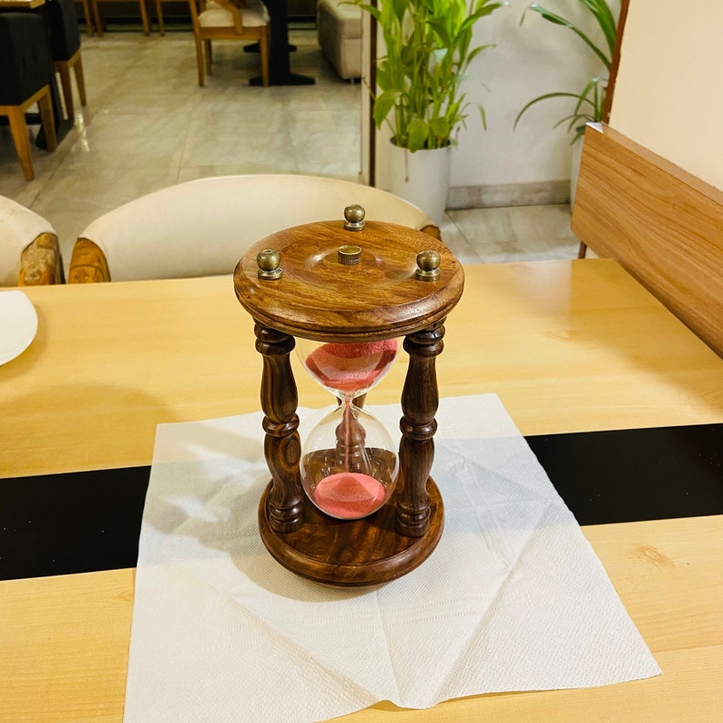 May include: A wooden hourglass with a dark brown finish and brass accents. The hourglass contains pink sand and is set on a wooden base. The hourglass is approximately 6 inches tall.