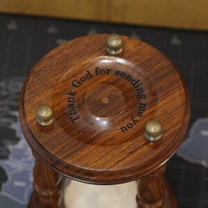 May include: Close-up of a wooden hourglass with a dark brown finish. The top has three brass-colored knobs and engraved text that reads "Thank God for sending me you."