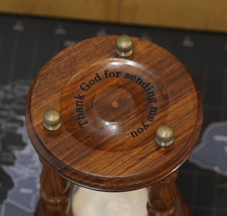 May include: Close-up of a wooden hourglass with a dark brown finish. The top has three brass-colored knobs and engraved text that reads "Thank God for sending me you."