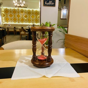 May include: A wooden hourglass with a dark brown finish and a clear glass bulb filled with pink sand. The hourglass is set on a white napkin on a wooden table. The hourglass is approximately 6 inches tall.