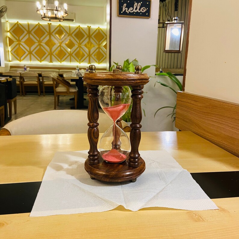 May include: A wooden hourglass with a dark brown finish and a clear glass bulb filled with pink sand. The hourglass is set on a white napkin on a wooden table. The hourglass is approximately 6 inches tall.
