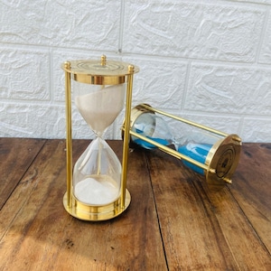 Könnte beinhalten: Zwei Sanduhren aus Messing. Eine Sanduhr hat weißen Sand und die andere blauen Sand. Beide Sanduhren haben eine Messingbasis und eine Messingspitze.