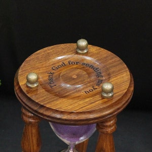 May include: A wooden hourglass with a dark brown finish and a purple sand filling. The top of the hourglass has the engraved text "Thank God for sending me you." Three brass-colored knobs are attached to the top. The hourglass is a decorative item.