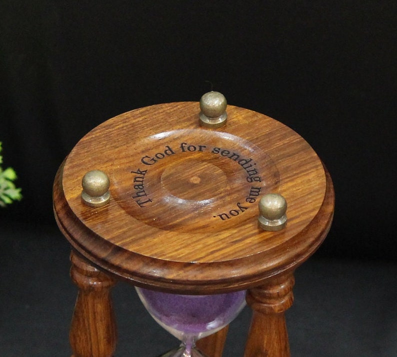 May include: A wooden hourglass with a dark brown finish and a purple sand filling. The top of the hourglass has the engraved text "Thank God for sending me you." Three brass-colored knobs are attached to the top. The hourglass is a decorative item.