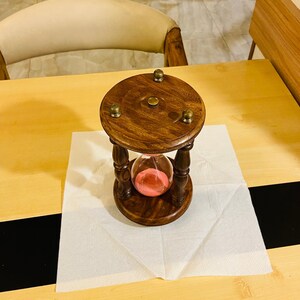 May include: A wooden hourglass with a dark brown finish, featuring a glass chamber filled with pink sand. The hourglass is supported by a turned wooden frame and sits on a white paper napkin. The top and bottom have brass accents.