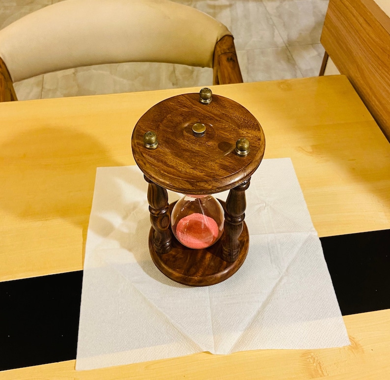 May include: A wooden hourglass with a dark brown finish, featuring a glass chamber filled with pink sand. The hourglass is supported by a turned wooden frame and sits on a white paper napkin. The top and bottom have brass accents.
