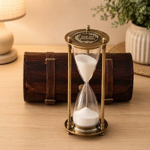 Könnte beinhalten: Eine Sanduhr mit Messingrahmen und transparenten Glaskammern, die mit weißem Sand gefüllt sind. Auf der Oberseite der Sanduhr steht der Text "LIVE THE LIFE YOU IMAGINED". Im Hintergrund befindet sich eine Holzkiste.