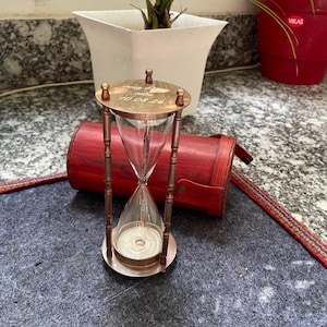 May include: A copper-toned hourglass with a clear glass bulb containing white sand. The top metal plate is engraved with the text "craig & caba" and "10.08.26". The hourglass features four slender vertical support pillars and a circular base.