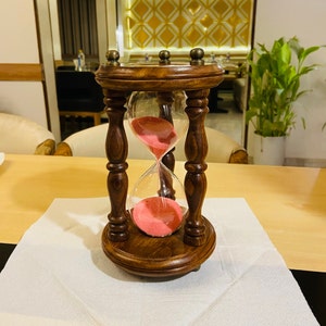 May include: A wooden hourglass with a dark brown finish. The hourglass has a clear glass chamber filled with pink sand. The hourglass is sitting on a white napkin on a light brown table.