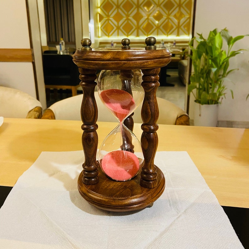 May include: A wooden hourglass with a dark brown finish. The hourglass has a clear glass chamber filled with pink sand. The hourglass is sitting on a white napkin on a light brown table.