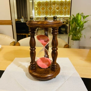 May include: A decorative hourglass with a dark brown wooden frame and base. The glass bulb contains pink sand. The hourglass is set on a white surface, with a light-colored wooden table in the background.